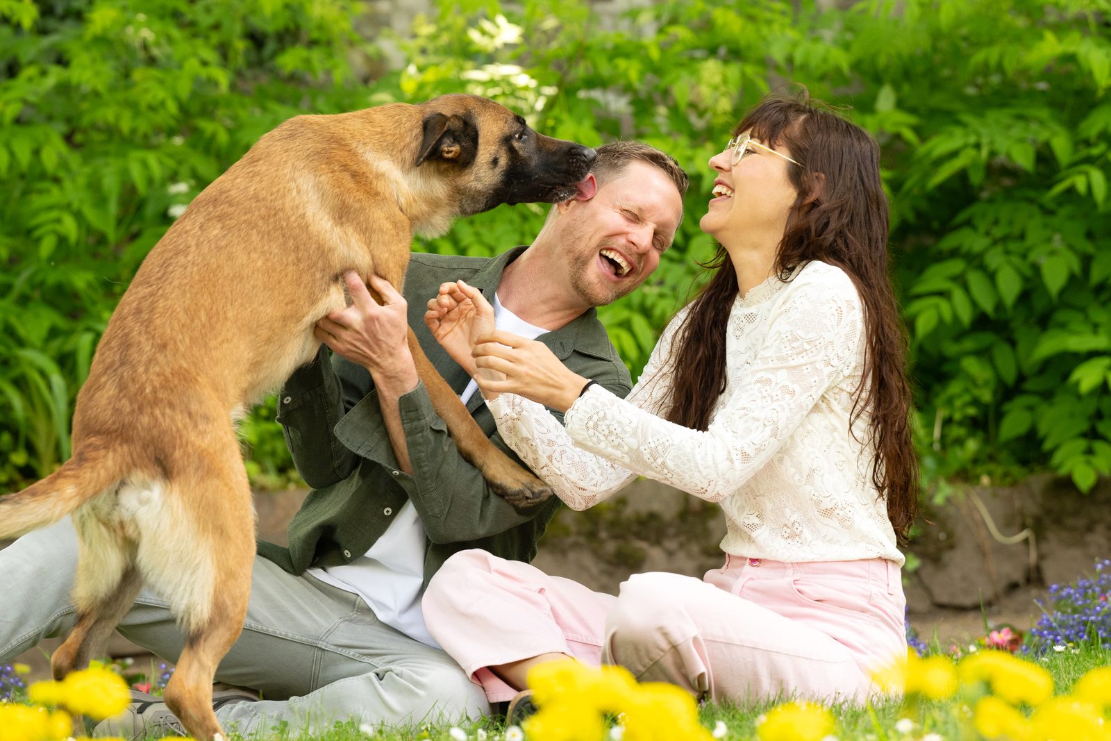 Clemens und Ronja lachen mit ihrer Schäferhündin Mali auf einer Blumenwiese